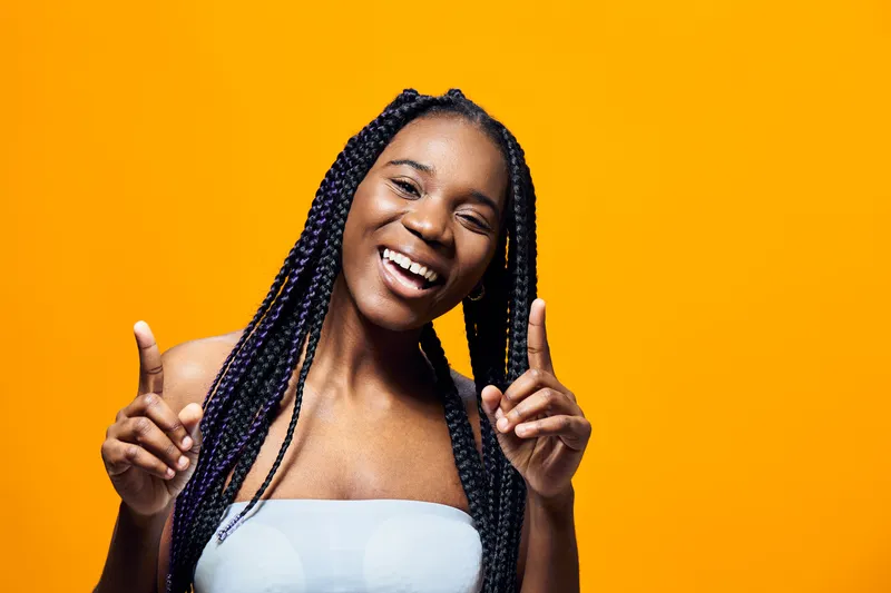 Happy young woman with braided hair against a bright yellow background, celebrating joy with playful gestures and a radiant smile, representing positivity and confidence