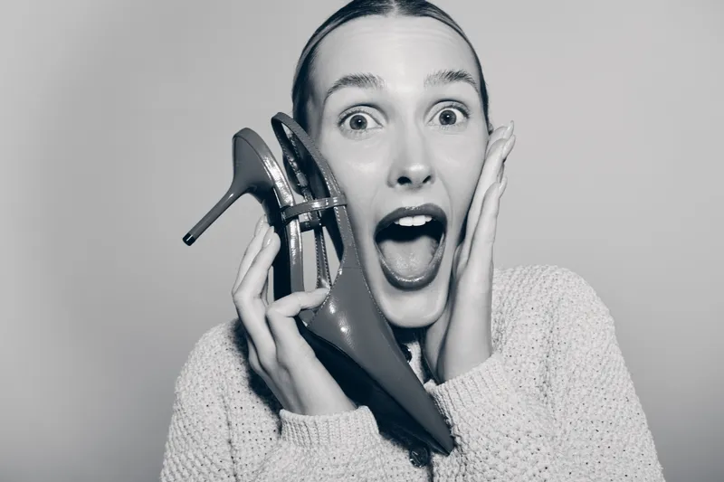 Surprised Caucasian woman with pale skin holding high heel shoe near face in studio. Concept of excitement and fashion.