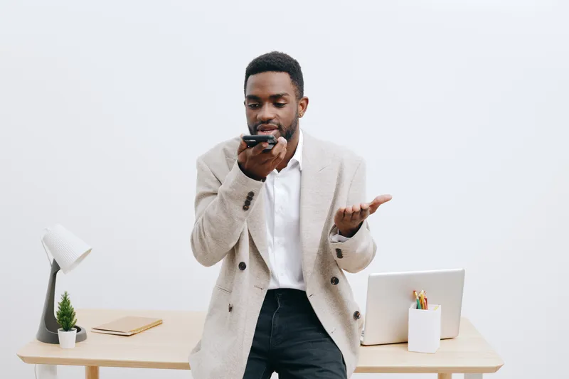 Professional young man with dark skin, wearing a beige cardigan and white shirt, standing in modern office, holding smartphone and gesturing with hand, working or talking, lifestyle concept, indoor sh
