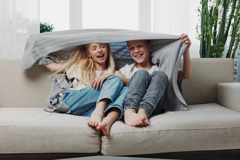 Two happy children sitting barefoot on sofa under a blanket, laughing and enjoying playful moment in bright living room. Concept of childhood joy and family comfort.