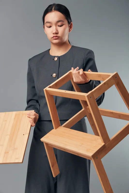 Woman holding wooden chair and table part in studio, furniture assembly or repair concept. Female with dark hair wearing jacket focused on construction and craftsmanship with tools or materials.