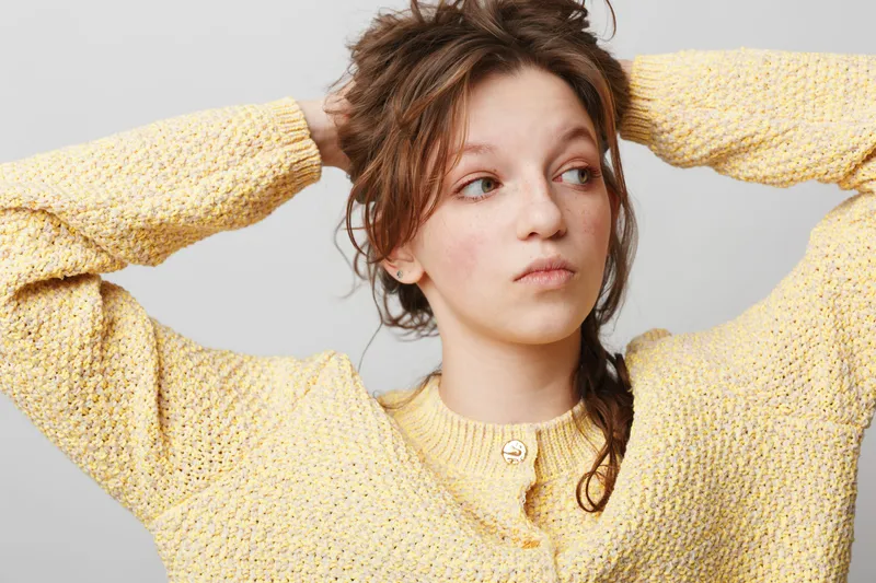 Young woman in yellow knitted sweater looks to side with thoughtful expression, hands behind head. Studio portrait on grey background. Concept of contemplation and casual fashion.