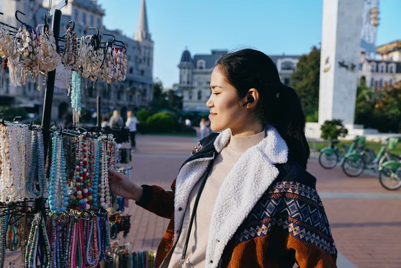 Woman in warm jacket browsing a bead jewelry stall at an outdoor market in a city square, selecting colorful accessories for style and gifting