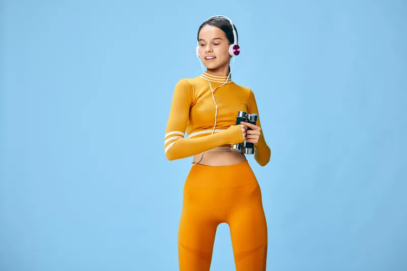 Fitness woman wearing orange athletic outfit, headphones, and holding a drink bottle against a blue studio backdrop. Active sports scene emphasizing energy, focus, and contemporary workout style for
