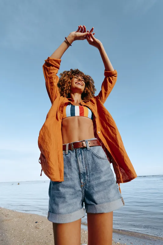 Happy young woman in orange shirt and striped bikini top enjoys sunny day at the beach with arms raised, smiling and feeling free near calm sea water.