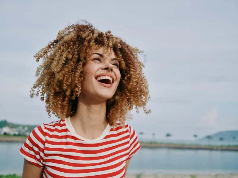 Joyful young woman with curly hair laughing against a bright sky and calm sea, wearing a red striped tee, capturing carefree summer energy and natural happiness.