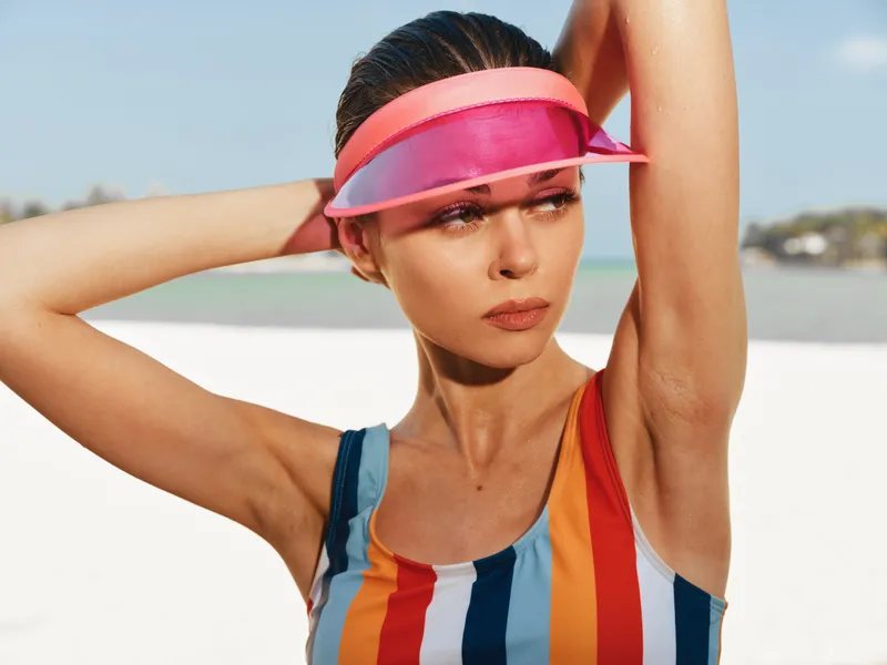 Portrait of a confident woman on a sunlit beach, wearing a striped swimsuit and a pink visor, posing with arms raised, vibrant colors and warm light.