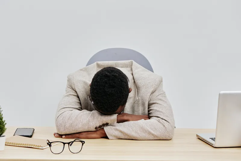 Businessman with dark hair hunched over desk resting head on arms exhaustion stress tiredness office workspace minimalistic background modern professional concept