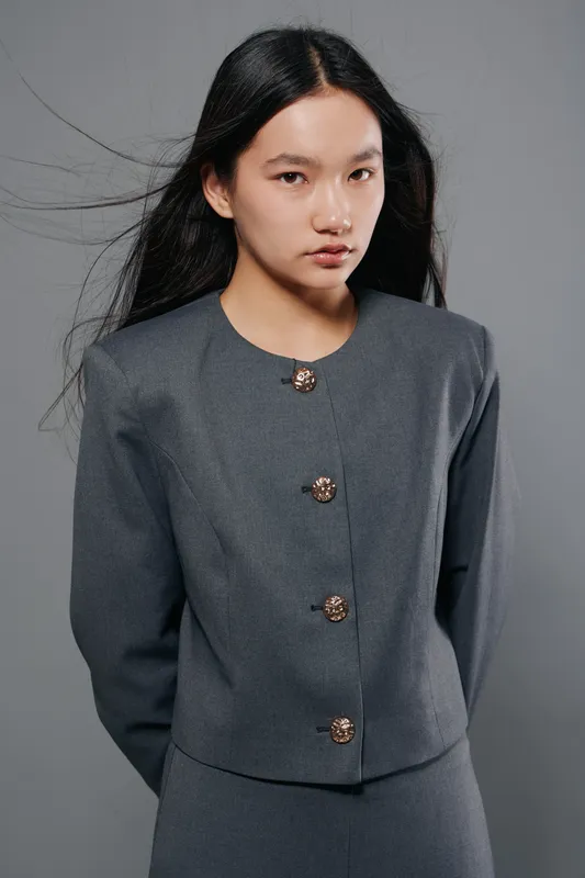 Woman with long black hair wearing gray formal jacket with gold buttons poses against plain gray background. Fashion model looking at camera with serious expression in studio setting.