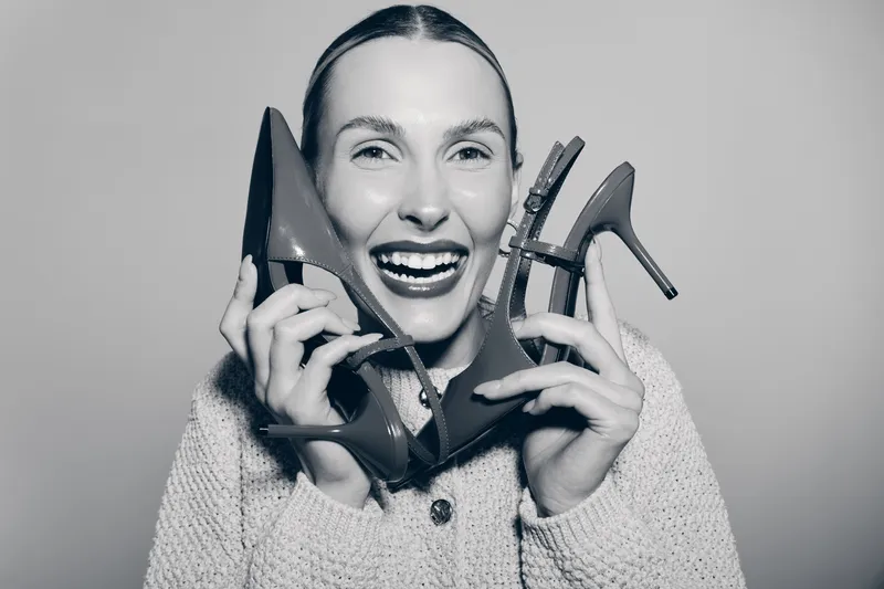 Happy Caucasian woman with light skin holding high heel shoes near face, smiling in studio. Concept of fashion, style, and footwear.