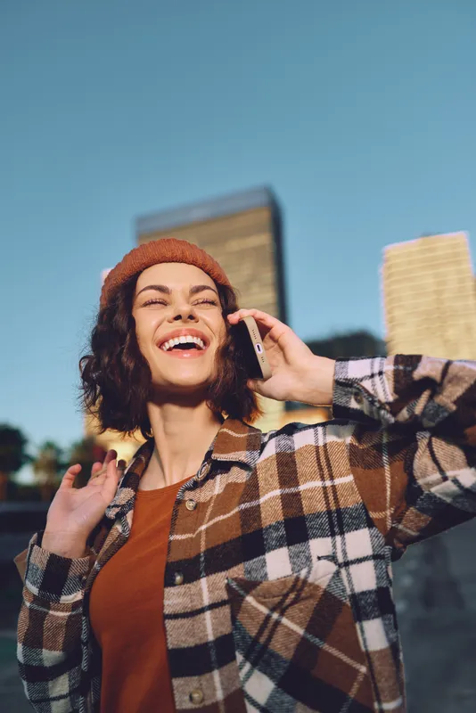 woman on phone with bright smile in city, candid lifestyle portrait bathed in golden glow, autumn beanie and plaid jacket, joyful authentic moment conveying mindfulness, emotional storytelling.