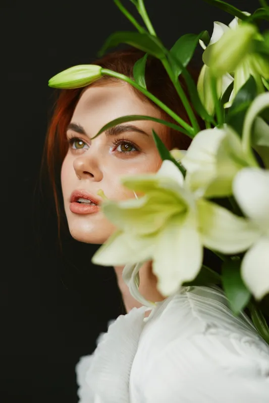 Portrait of a model with green eyes and red hair surrounded by white lilies, demonstrating elegance and natural beauty in a dark, minimalist setting