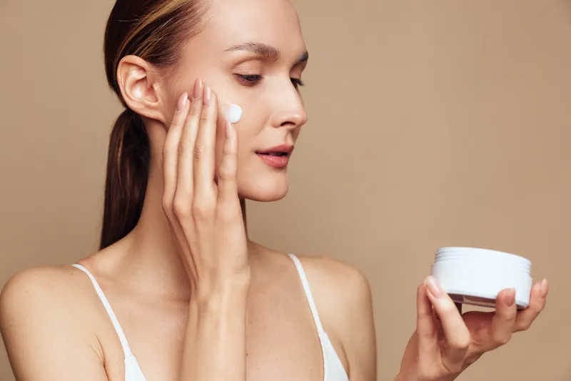 Woman skincare face cream beauty spa caring wellness, young woman applies moisturizer, holding skincare jar, smooth natural skin, isolated on beige studio background