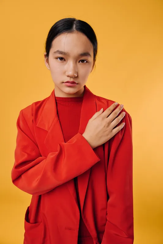Young woman in red jacket with serious expression posing against orange background. Fashion portrait with minimal makeup and neat hairstyle in studio. Stylish female model looking at camera.