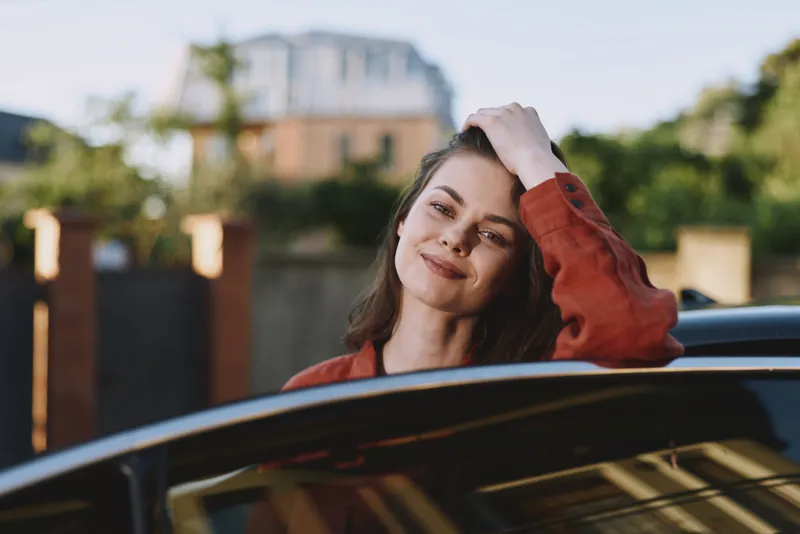 Cheerful woman in a car, outdoor lifestyle moment, warm sun, relaxed pose, soft breeze, natural colors, urban background, candid portrait