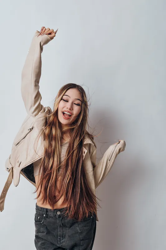 Young woman with long hair wearing jacket and black pants dancing and smiling with closed eyes against a light background. Casual fashion, joyful expression and relaxed mood.