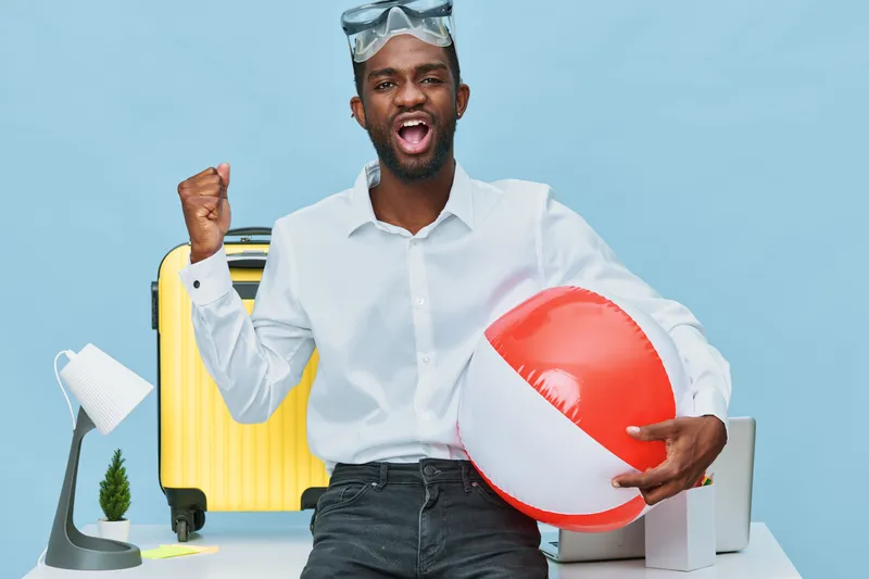 Cheerful young man with dark skin, wearing white shirt, holding beach ball and smiling against light blue background. Office supplies, yellow suitcase, lamp, and laptop are visible, creating a fun sum