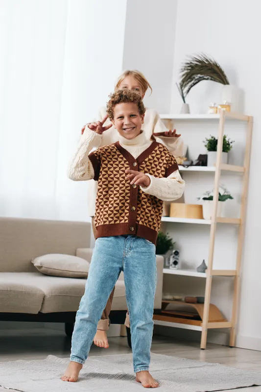 Two joyful children play indoors in a cozy living room, one pointing at the camera while the other hugs them from behind, wearing warm knitwear and relaxed denim.