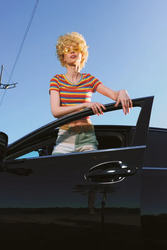 A stylish woman with curly blonde hair leans from a sleek car window, wearing a striped tee under bright sun. Retro lifestyle vibe and bold color palette capture a carefree road trip.