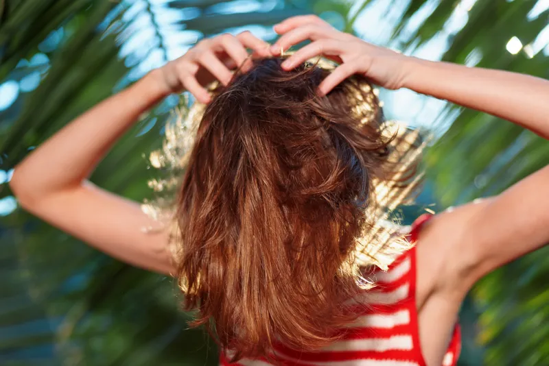 An individual with sunlit hair in a red striped top stands outdoors, hands in their hair, surrounded by tropical foliage, conveying a carefree, energetic summer mood.