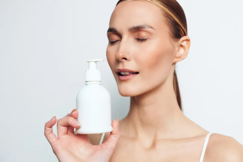 Young woman with clear skin holds white cosmetic bottle with pump dispenser close to face eyes closed isolated on white background beauty skincare concept.