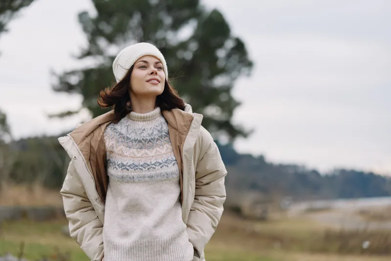 Woman standing outdoors in a cozy beige jacket and knit sweater, beanie, calm countryside setting with soft natural light, showcasing relaxed style and autumn mood, simple composition and tranquil