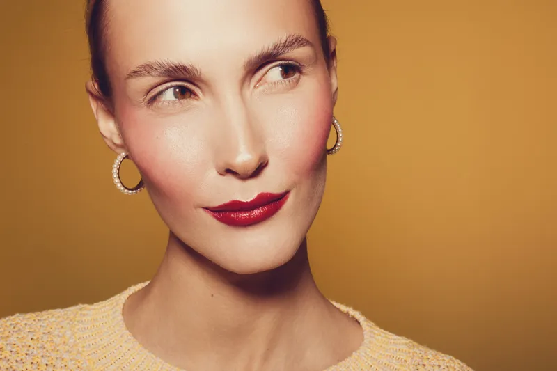 Close-up portrait of a woman with red lipstick and hoop earrings looking sideways on a warm beige background. Concept of beauty, makeup, fashion, and elegance.