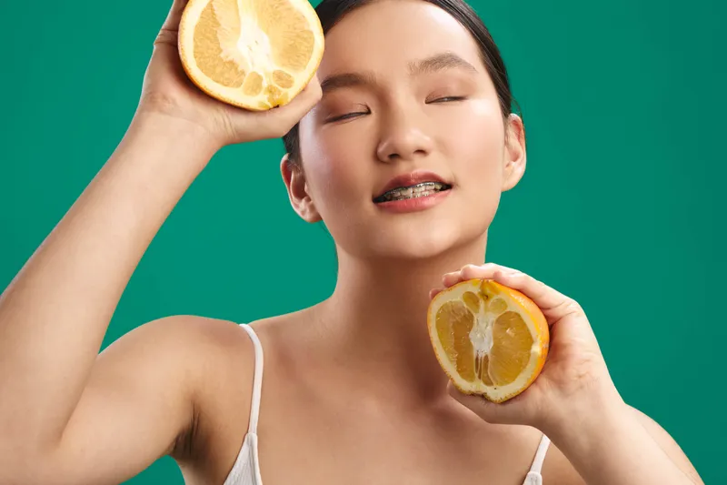 Asian woman with smooth skin smiles gently as she holds two lemon halves near her cheeks, vibrant green background framing a fresh, playful portrait that exudes energy, brightness, and wellness.