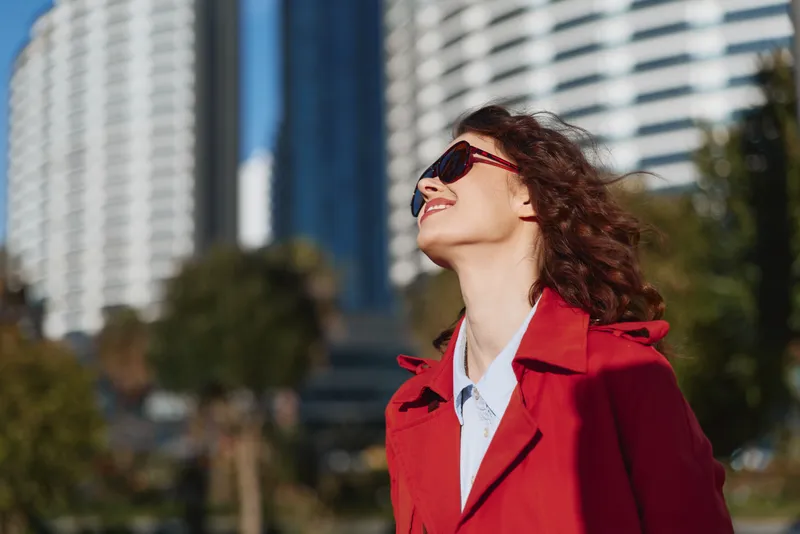 Young woman with curly hair enjoys a sunny day outdoors wearing a red coat and sunglasses with a cityscape background.