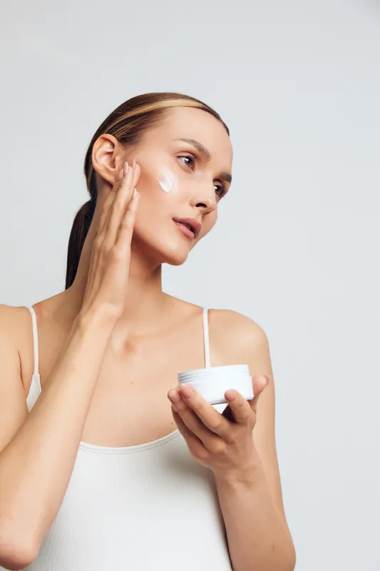 Young woman applies moisturizing cream on face with fingers holding white jar skin care routine natural beauty isolated on light background healthy skin concept