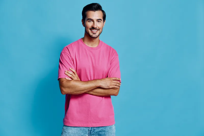 Smiling man wearing pink t-shirt stands confidently with crossed arms against a solid blue background, showcasing a cheerful lifestyle concept.