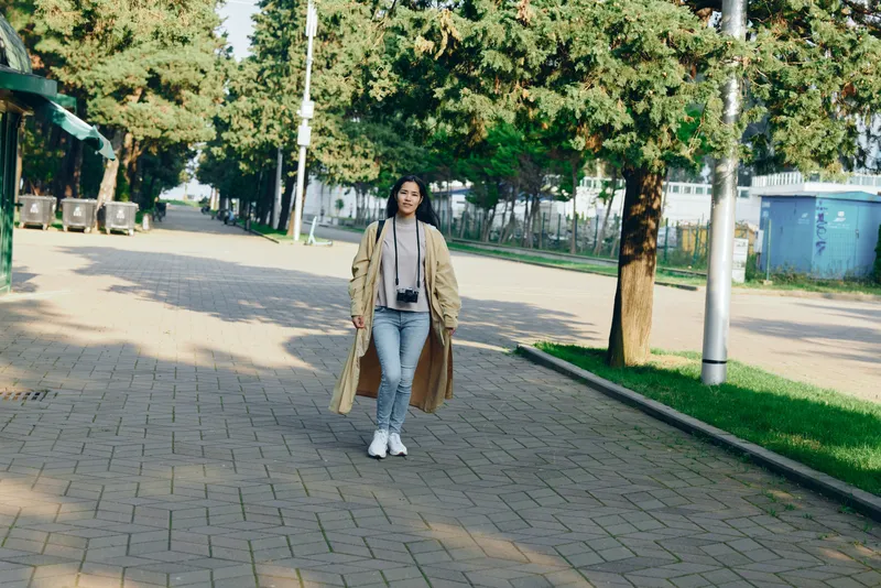 Woman walking in a park wearing a long beige coat, jeans and sneakers along a sunlit pathway. Casual urban style, relaxed mood, outdoor scenery, greenery, calm city stroll