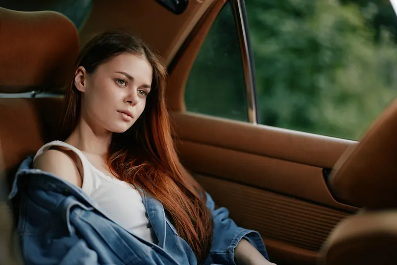 Pensive young woman with long red hair sitting in a car, wearing a denim jacket, looking thoughtfully out the window. Emotional portrait captured in natural light.