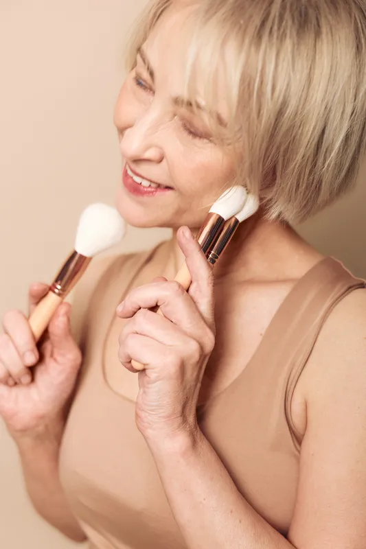 Happy middle aged woman with short blonde hair gently applying powder with two makeup brushes. Soft beige background and natural light create warm skincare and beauty concept.