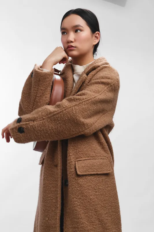 Asian woman stands in a studio setting, wearing a warm brown coat and holding a guitar case, eyes calm and thoughtful, creating a quiet, creative mood.
