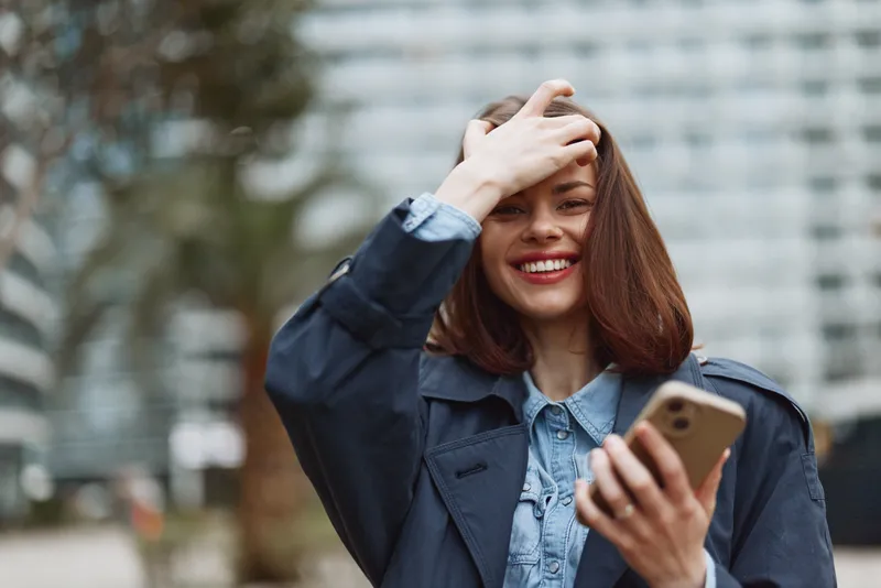 Smiling woman outdoors holds a smartphone, hand in hair. Casual fashion in an urban setting, candid portrait showing everyday connection and ease.