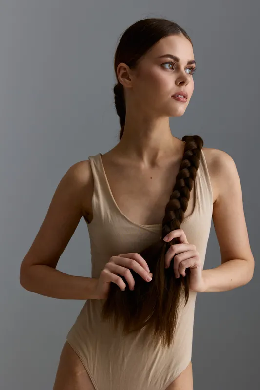 young woman with long braided hair wearing a beige sleeveless bodysuit, standing against a minimalist gray background, natural light, thoughtful expression
