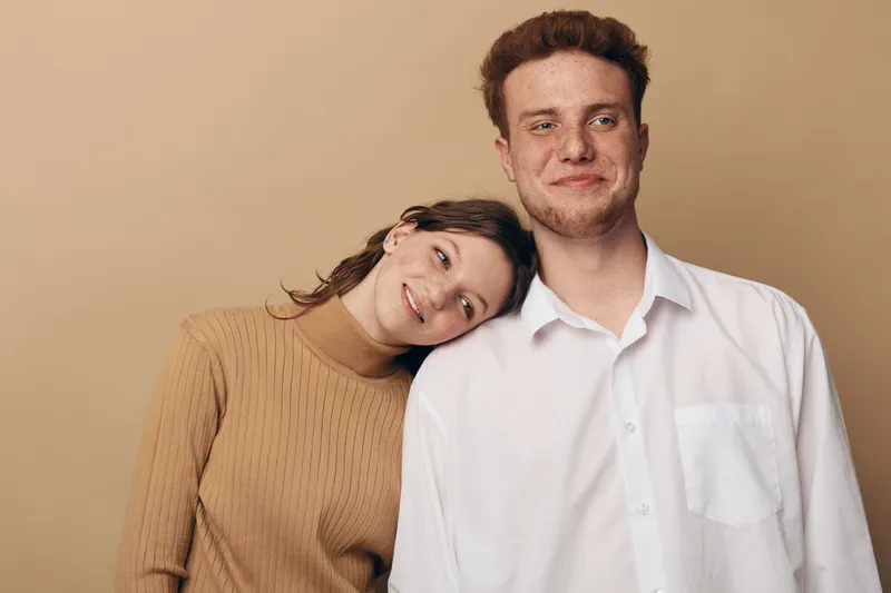 Young couple with woman leaning on man shoulder smiling gently. Casual clothing and warm beige background create cozy, intimate atmosphere. Concept of love, support, and togetherness.
