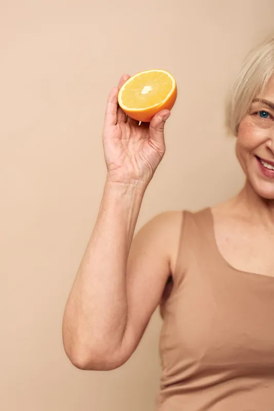 Smiling mature woman holds half of a fresh orange close to her face against beige background. Concept of healthy lifestyle, natural vitamins, nutrition, wellness, and positive mood.