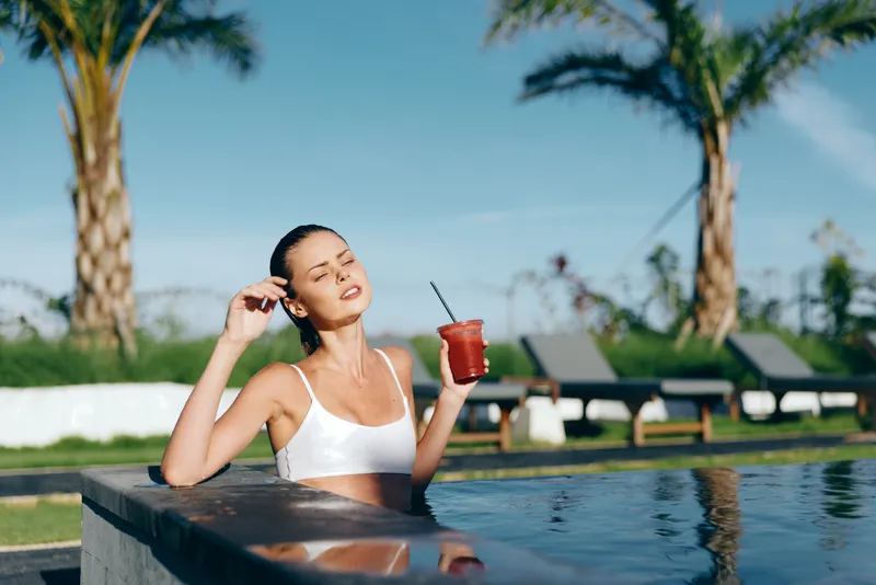 Young woman in white swimsuit relaxing in outdoor pool holding a red drink with straw, enjoying sunny day with palm trees and lounge chairs in the background, lifestyle concept.
