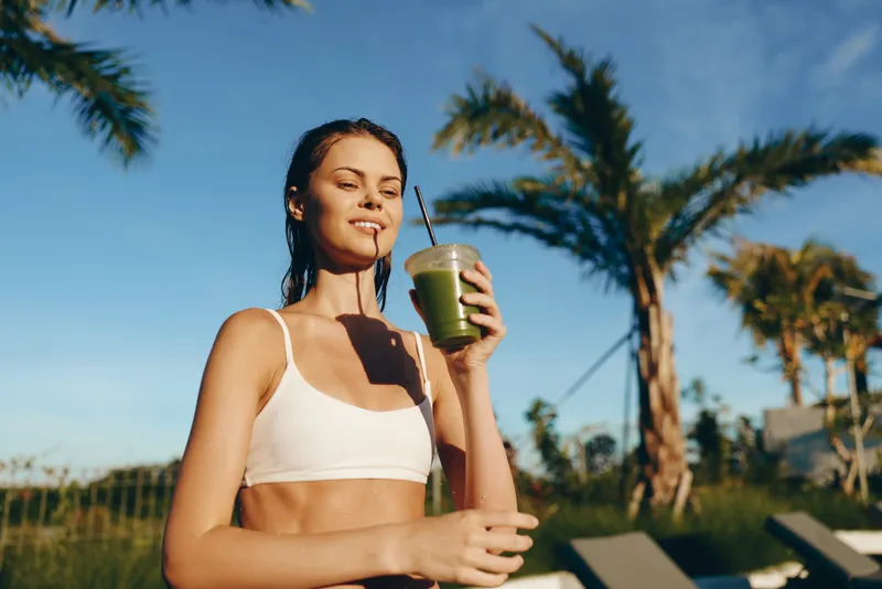 Active woman enjoys a refreshing smoothie after a workout in outdoor sun. Palm trees frame a bright scene, conveying fitness, health, lifestyle, summer, relaxation and tropical vibes.