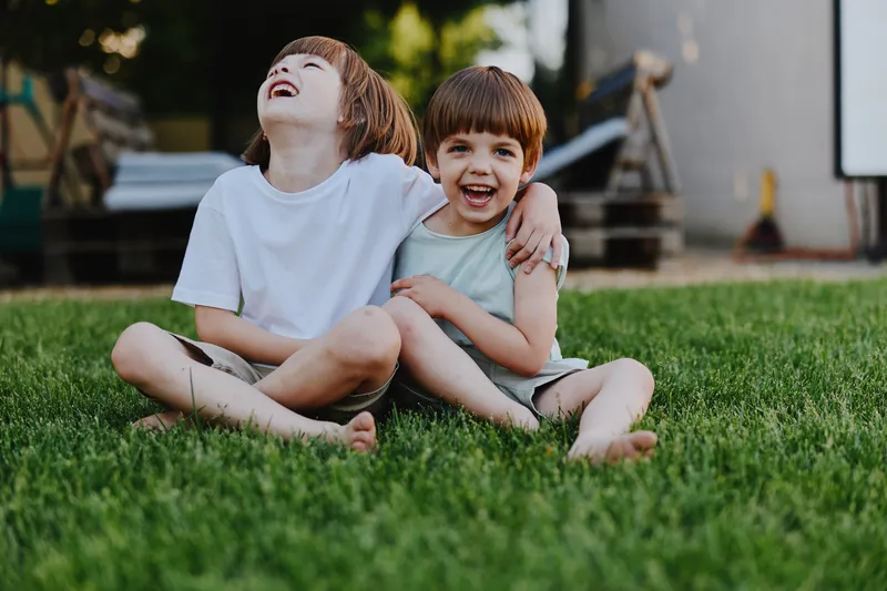 Two happy boys laughing together while sitting on the grass, enjoying a sunny day. Their playful expressions capture the innocence of childhood in a natural setting.