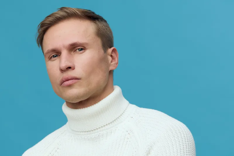 Portrait thoughtful young caucasian man wearing white turtleneck sweater isolated on blue background digital detox concept