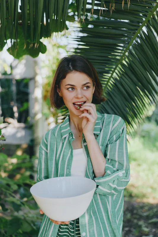 Woman outdoors enjoying a snack, holding a white bowl beneath tropical palm leaves, wearing a green striped shirt in natural light.