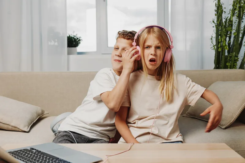 Two children sitting on sofa with headphones, boy annoys girl while she listens to music at home. Concept of sibling interaction and childhood emotions.