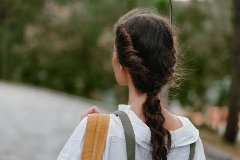 A young woman with long braided hair walking outdoors, wearing a casual white shirt, showcasing a serene expression amidst a lush green background, symbolizing relaxation and connection with nature