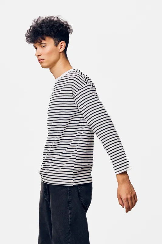 young man wearing striped long sleeve shirt and black pants standing sideways against plain white background with thoughtful expression and casual style