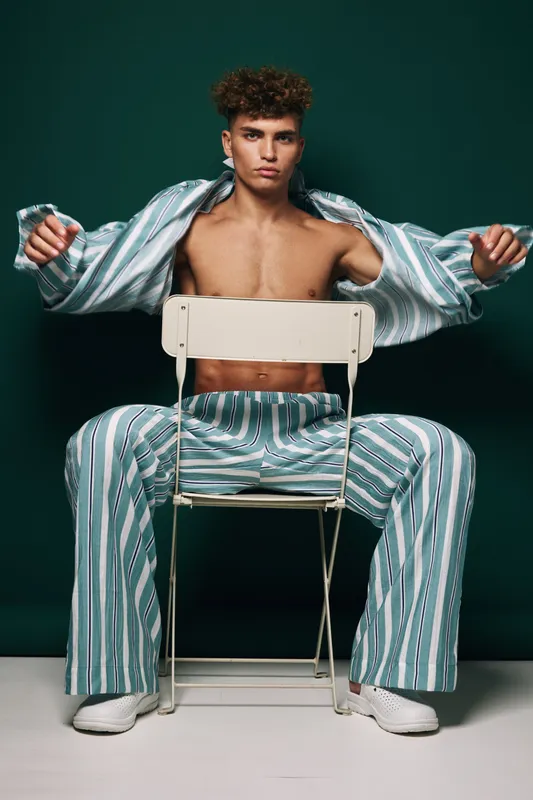 Young man with curly hair sitting on chair backward wearing striped pants and open shirt showing bare chest in studio with dark green background and white shoes.