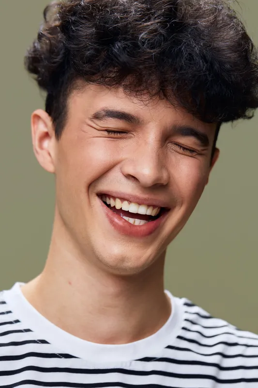 happy man laughing with closed eyes in striped shirt, curly hair, joyful expression, isolated on green background, young adult male portrait, casual style