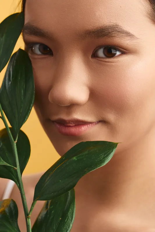 Asian woman is captured in a closeup portrait with lush green leaves in a warm studio setting. She gazes softly, radiating calm, beauty, and natural freshness.